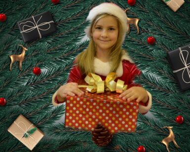 Zbliżenie. Dziewczynka w czapce Mikołaja trzyma prezent. W tle gałęzie choinki, prezenty i renifery.