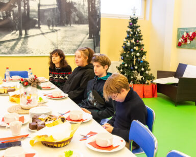 Sala. Czterech młodych ludzi siedzi przy stole wigilijnym. W tle choinka z ozdobami świątecznymi, pod choinką prezenty.