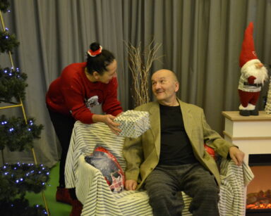 Zbliżenie. Kobieta trzyma prezent, patrzy na mężczyznę. Senior siedzi na kanapie. Obok kominek i świąteczna choinka.