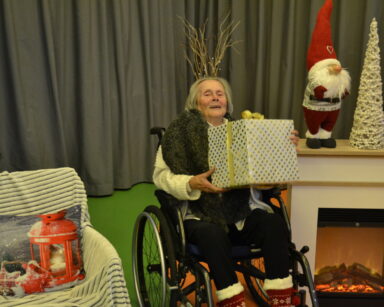 Zbliżenie. Kobieta na wózku trzyma w ręku duży prezent. Obok kominek na nim choinka i dwa Mikołaje.