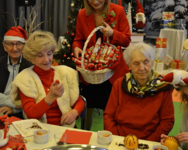 Sala. Kobieta z koszem słodyczy stoi za seniorami. Przy stole siedzą seniorzy, na stole leżą pomarańcze z goździkami.