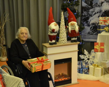 Zbliżenie. Kobieta na wózku trzyma prezent. Obok kominek na nim ozdoby świąteczne. W tle choinka i prezenty.
