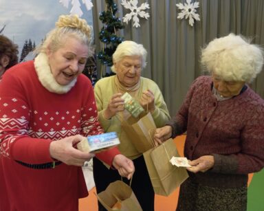 Zbliżenie. Trzy uśmiechnięte seniorki rozpakowują prezenty. W tle zimowy krajobraz i świąteczne choinki.