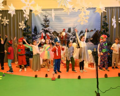 Sala. Scena. Grupa maluchów w kolorowych strojach wykonuje przedstawienie. W tle zimowa sceneria, choinki, szyszki.
