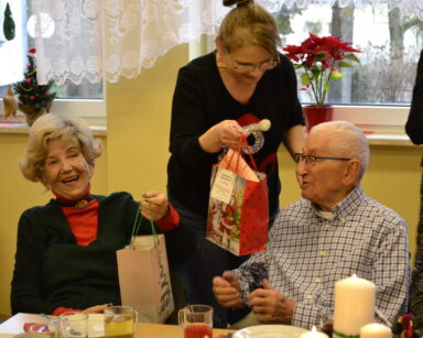 Sala. Uśmiechnięci seniorzy siedzą przy stole. obok stoi kobieta i trzyma w ręku prezent. Na stole wigilijne potrawy.