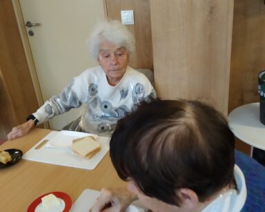 Zbliżenie. Dwie seniorki siedzą przy stole. Jedna smaruje kromkę chleba masłem, druga seniorka się przygląda.