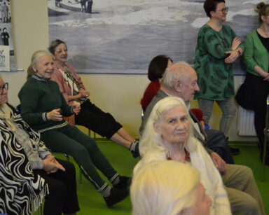 Zbliżenie. Grupa seniorów ogląda przedstawienie. Pod ścianą stoją dwie kobiety i uśmiechają się.