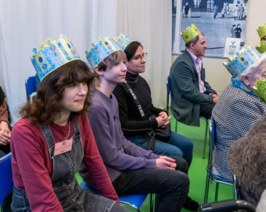 Sala. Grupa ludzi w różnym wieku siedzi na krzesłach. Na głowie mają korony w kolorach niebiesko żółtym.