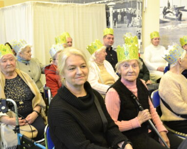 Sala. Grupa osób ma na głowie korony w kolorze niebiesko żółtym. Jedna z seniorek uśmiechnięta pozuje do zdjęcia.