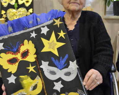 Zbliżenie. Seniorka pozuje do zdjęcia w rękach trzyma tablicę z kolorowymi maskami karnawałowymi i gwiazdkami.