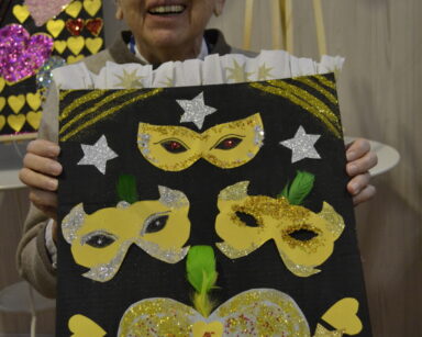 Zbliżenie. Seniorka trzyma w dłoniach tablicę z kolorowymi maskami i sercem. Kobieta pozuje do zdjęcia.