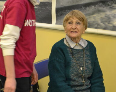 Zbliżenie. Uśmiechnięta seniorka siedzi na krześle i patrzy na wolontariusza. W ręku trzyma kulę do kręgli.