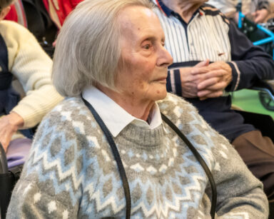Zbliżenie. Seniorka w kremowym swetrze we wzorki jasnej koszuli ogląda przedstawienie. Obok siedzą seniorzy .