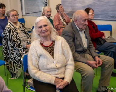 Zbliżenie. Seniorka w kremowym swetrze i złotej koszulce pozuje do zdjęcia. Obok siedzą seniorzy i oglądają występ.