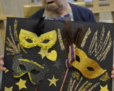 Zbliżenie. Seniorka pozuje do zdjęcia. W dłoniach trzyma tablicę ze złotymi maskami, obok przyklejone serduszka.