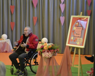 Zbliżenie. Senior na wózku śpiewa. Unosi dłonie. Obok napis Dzień Dziadka. W tle szara kotara i czerwone zwisające serca.