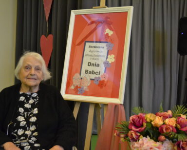 Zbliżenie. Seniorka pozuje do zdjęcia przy plakacie z napisem Serdeczne życzenia z okazji Dnia Babci.
