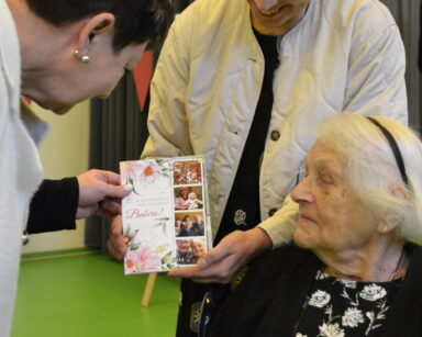 Zbliżenie. Dwie kobiety trzymają kartkę z napisem Wszystkiego najlepszego Babciu. Obok siedzi seniorka i patrzy na kartkę.