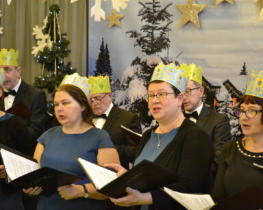 Zbliżenie. Grupa osób w koronach na głowie trzyma w rękach śpiewniki. W tle ozdoby świąteczne.