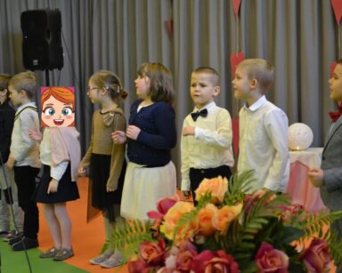 Zbliżenie. Ośmioro przedszkolaków stoi na scenie. Obok mikrofon, głośnik, bukiet kwiatów. Szara kotara i serduszka.