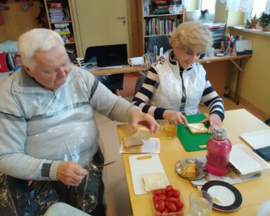 Sala. Para seniorów siedzi przy stole i smaruje masłem kromki chleba. Na stole ciasto, pomidorki, różowa butelka i tacki.