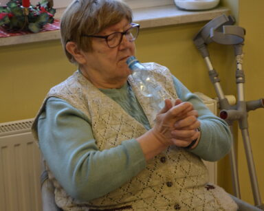Zbliżenie. Seniorka w jasnym swetrze i szarej kamizelce trzyma w dłoniach zgniecioną plastikową butelkę.