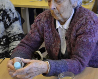 Zbliżenie. Seniorka trzyma w dłoni zgniecioną butelkę z plastiku. Przed kobietą na stoliku kawa w szklance.