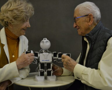 Zbliżenie. Para seniorów trzyma za rączki robota. Seniorzy uśmiechają się. Robot stoi na stoliku.