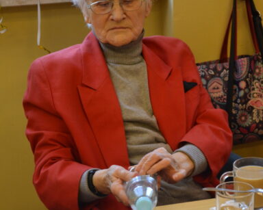 Zbliżenie. Seniorka w czerwonej marynarce siedzi przy stole. Kobieta patrzy na butelkę plastikową, którą trzyma w dłoni.