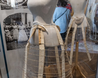 Zbliżenie. Dwa manekiny, na nich konstrukcja z drutu pod długą suknię. W tle zdjęcia kobiet ubierających stelaż pod suknię.