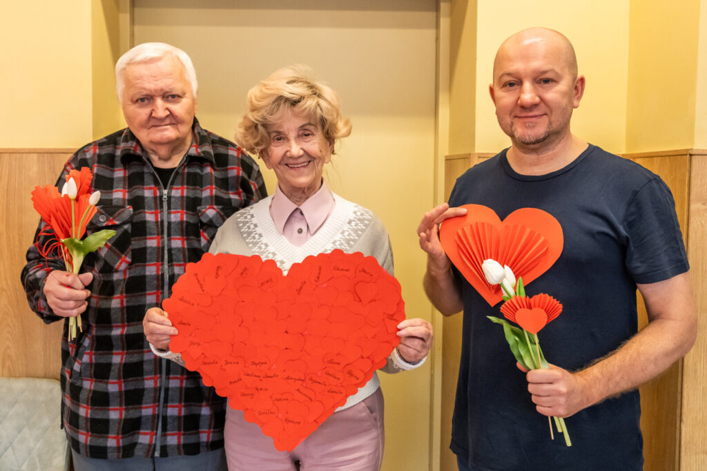 Trzy osoby pozują do zdjęcia. Dwóch mężczyzn trzyma białe tulipany i serduszka. Kobieta trzyma duże serce z imionami.