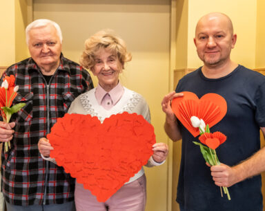 Trzy osoby pozują do zdjęcia. Dwóch mężczyzn trzyma białe tulipany i serduszka. Kobieta trzyma duże serce z imionami.