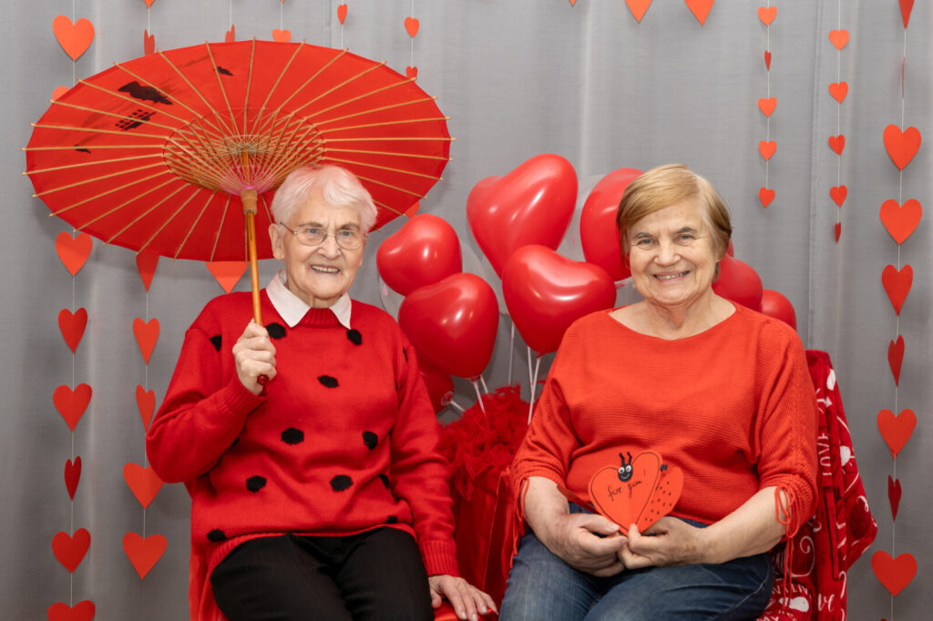 Zbliżenie. Dwie seniorki pozują do zdjęcia. Kobiety ubrane są w czerwone swetry. W tle czerwone balony, serduszka, parasol.