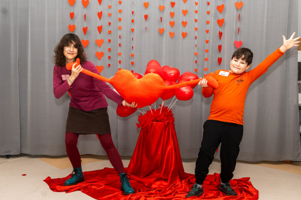 Scena. Para dzieci pozuje do sesji walentynkowej. W dłoniach trzymają serce z napisem "Kocham Cię." W tle balony serca.
