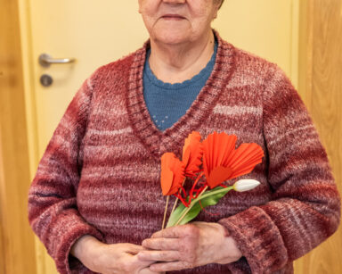 Zbliżenie. Seniorka w swetrze w pasy pozuje do zdjęcia. W dłoniach trzyma tulipana i kilka serc na patyczkach.