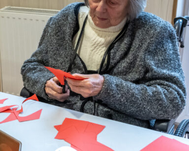 Zbliżenie. Seniorka siedzi przy stole i wycina z czerwonej kartki serca. Na stole nożyczki, kartki.