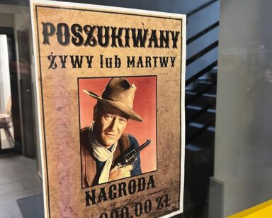 Zbliżenie. Na szybie plakat z kowbojem. To aktor John Wayne. Na plakacie wyznaczona nagroda za żywego lub martwego.