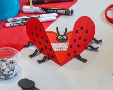 Stół. Na stole kolorowe oczka w pojemniku, pisaki, nożyczki i kartka w kształcie biedronki i serca z wierszykiem w środku.