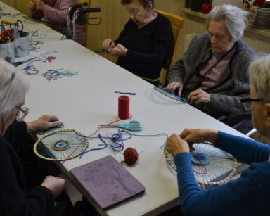 Sala. Stół. Seniorki przy stole przekładają włóczki przez koło. Tworzą mandale. Na stole kłębki włóczek, nożyce i nici.