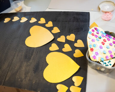 Zbliżenie. Na stole czarny karton a na nim żółte serca. W pudełku obok złote serca z kolorowymi kwiatkami.