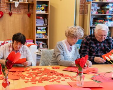 Sala. Stół. Przy stole trzech seniorów wycina czerwone serduszka. Na stole czerwony papier, serca i białe tulipany.