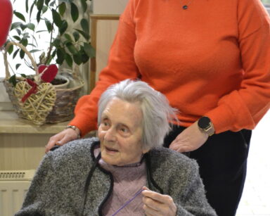 Sala. Kobieta w pomarańczowym swetrze stoi przy siedzącej seniorce. Seniorka w ręce trzyma włóczkę. Na stole leży kółko.