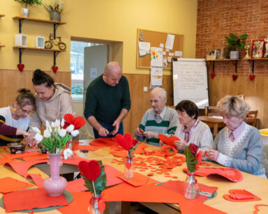 Sala. Grupa osób siedzi przy stole i wycina serduszka. Na stole czerwone kartki, białe tulipany. W tle ozdoby na ścianach
