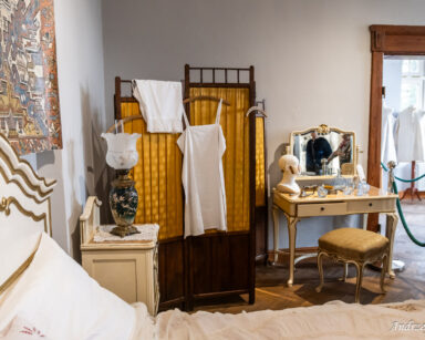 Pokój. Szare ściany, drewniany parawan z żółtym materiałem, toaletka, stolik z lampą, łóżko. Na toaletce flakony i lustro.