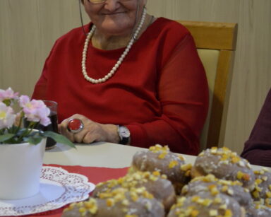 Zbliżenie. Seniorka w czerwonej sukience i koralach siedzi przy stole. Na stole stoją kwiatki i talerz z pączkami.