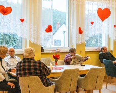 Sala. Grupa seniorów siedzi przy stołach. W tle przy oknach z sufitu na sznurku zwisają czerwone serca.