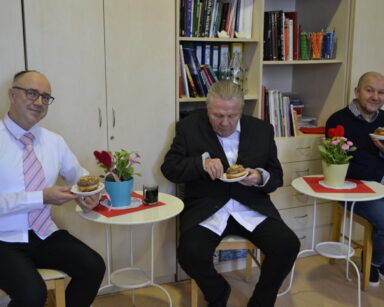 Zbliżenie. Trzech mężczyzn przy stolikach trzyma w ręku talerzyk z pączkiem. Na stolikach kwiatki, kawa, czerwone serwetki.