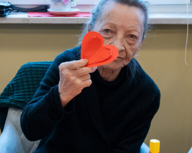 Zbliżenie. Seniorka pozuje do zdjęcia. W dłoni trzyma ozdobne czerwone serce. Na stole przed nią dwa czerwone serca.