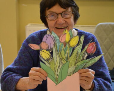 Zbliżenie. Uśmiechnięta seniorka siedzi przy stole i trzyma w rękach bukiet wyciętych i pokolorowanych tulipanów.