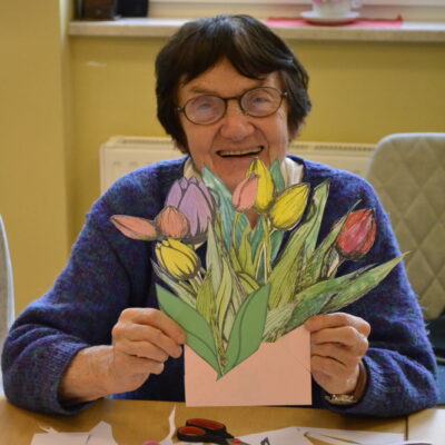 Zbliżenie. Uśmiechnięta seniorka siedzi przy stole i trzyma w rękach bukiet wyciętych i pokolorowanych tulipanów.